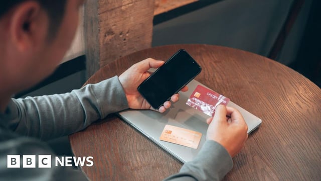 Man holds a credit card and a phone with another bank card and a laptop on the table in front of him.