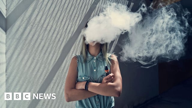 Young lady wearing blue demin shirt and jeans, She has her arms crossed and is holding a disposable vape, with a plume of smoke obscuring her face.