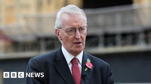 Hilary Benn has white hair and wears thin black glasses. He is wearing a black suit with a red tie. He has a red poppy on the left side of his blazer.
