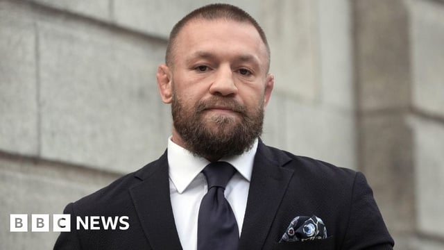 Conor McGregor is wearing a black suit with a white shirt and a black tie. He's outside a courthouse. There's a patterned pocket squire in his jacket pocket.