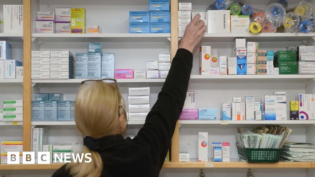 The photo shows a woman with blonde hair tied in a low ponytail, with glasses on her head, wearing a black jumper. She is facing away from the camera and reaching onto the top shelf of a wall full of different medications.