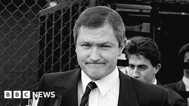 A black and white still of Pat Finucane, who is wearing a suit jacket, a shirt and striped tie. He is walking out of a court building while holding a folder and files.