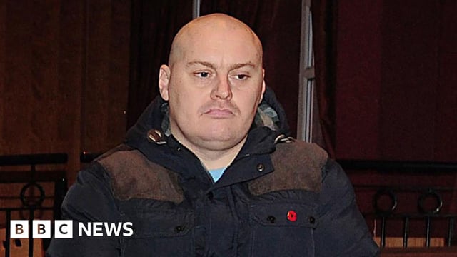 Ian Ogle is seen wearing a navy coat with a poppy pin.
He is bald, with a light stubble and frowning.