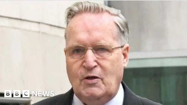 Close-up of Paul Dunleavy outside court. He has grey-ish hair, wearing glasses, black blazer and white shirt.