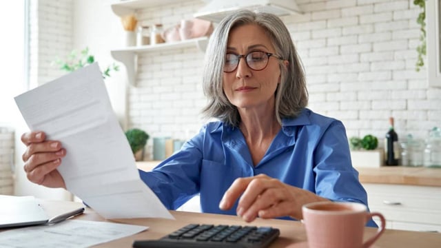 A woman is seen sitting at a kitchen table with a calculator looking at the Social Security benefits she will receive in retirement.
