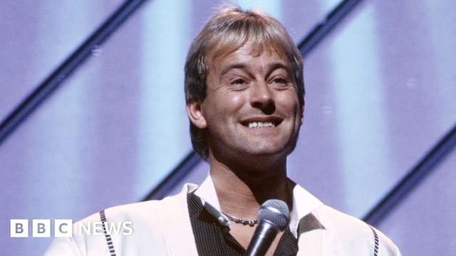 Duncan Norvelle at London Palladium in 1987 in a white shirt holding a microphone