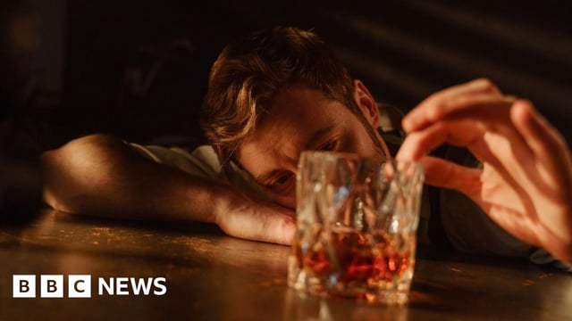 A man with brown hair and a beard leans his head on his arm on a table and gazes into a glass of whisky while running his index finger around the rim of the glass.