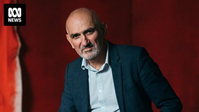 Australian singer-songwriter Paul Kelly poses in front of a red backdrop.