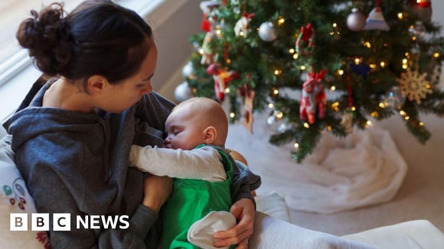 A woman sits on a sofa next to a Christmas tree and breastfeeds her baby