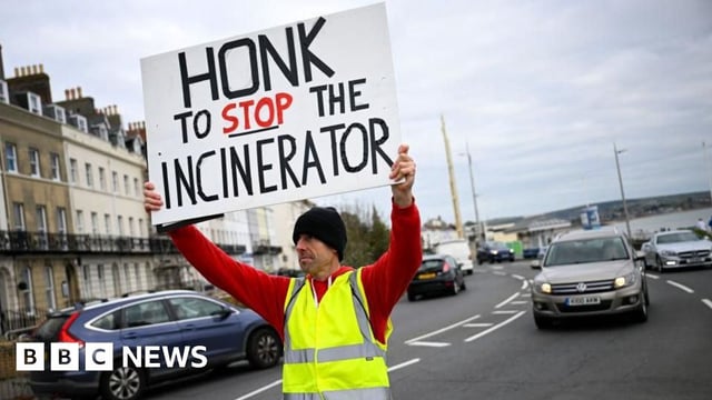 A man in a hi-vis jacket holds a sign that reads: Honk to stop the incinerator. The word stop is underlined. He is standing in the middle of the road facing the camera, with a car behind him, and a row of houses to his right.