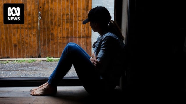 A young woman is silhouetted by a window, wearing a cap and hugging herself.