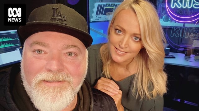 A man with a grey beard and black cap and a blonde woman resting her hand on his shoulder smile in a radio studio.