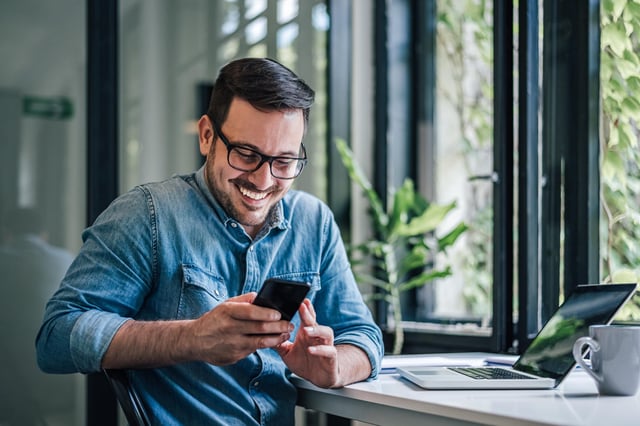 In this stock image, a man sitting at his computer with a cup of coffee smiles while looking at his mobile phone.