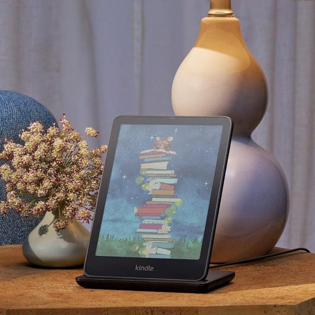a kindle colorsoft e-reader sits on a side table on a stand with a lamp sitting behind it