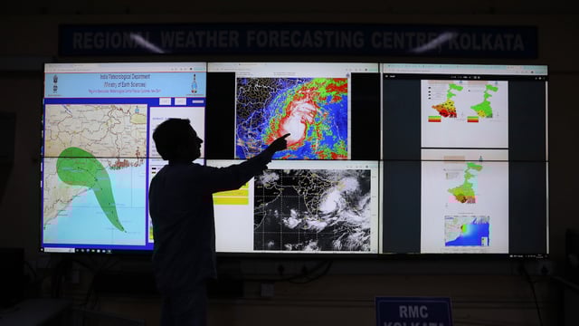 A meteorologist seen in silhouette points toward a large screen displaying the position of a cyclone.