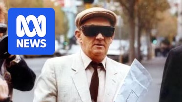 A man with a white cap and black sunglasses walks between a TV cameraman and another man dressed in black.