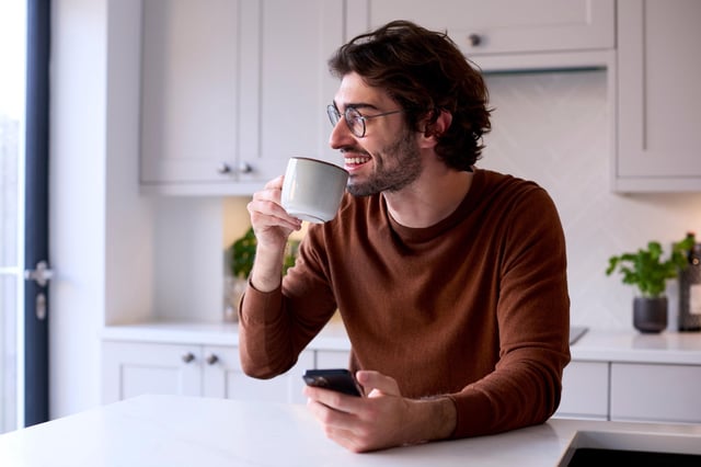 A man relaxes at home in his kitchen while holding a coffee and his mobile phone.