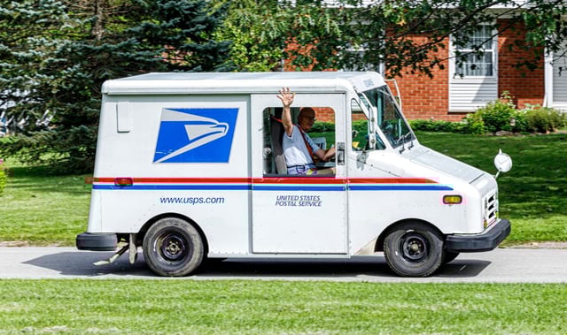 A local postal worker driving an official government United States Postal Service (USPS) mail delivery vehicle van is waving out the window to an acquaintance as he delivers mail in this residential district neighborhood located in the town of Penfield, NY, on September 1, 2017.