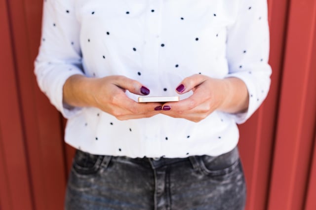 In this stock image, a woman with painted nails wearing a white polka dot shirt types on her mobile phone.