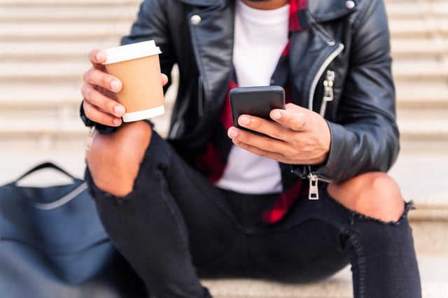 A man wearing a leather jacket and ripped black jeans holds a mobile phone in one hand and a coffee in the other.