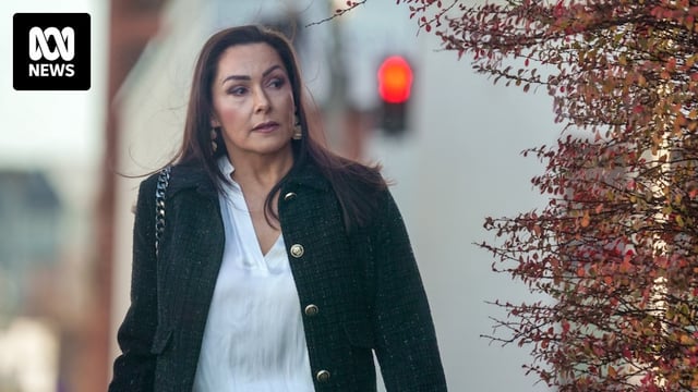 A woman with windswept, dark hair wearing a black coat and white blouse turns a street corner with autumn foliage.