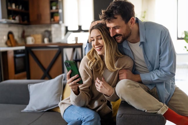 In this stock image, a cozy couple embraces while looking at a mobile phone at home.