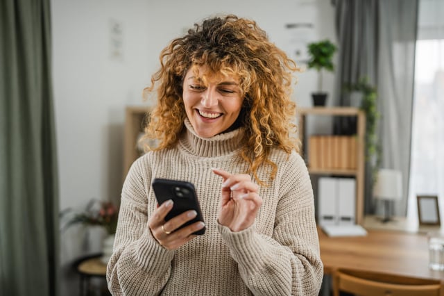 In this stock image, a woman with curly hair smiles while using her mobile phone at home.