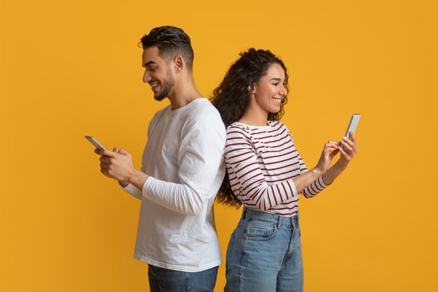 In this stock image, a young couple stands back-to-back while looking at their mobile phones against a yellow background.