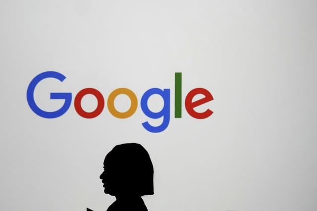 FILE - A woman walks by a giant screen with a logo at an event at the Paris Google Lab on the sidelines of the AI Action Summit in Paris, Sunday, Feb. 9, 2025. (AP Photo/Thibault Camus, file)