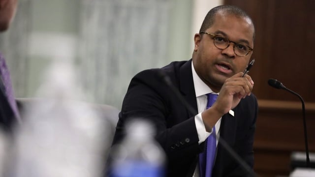 FCC Commissioner Geoffrey Starks sits at a table and speaks into a microphone while holding a pen in his right hand while testifying during a Senate hearing