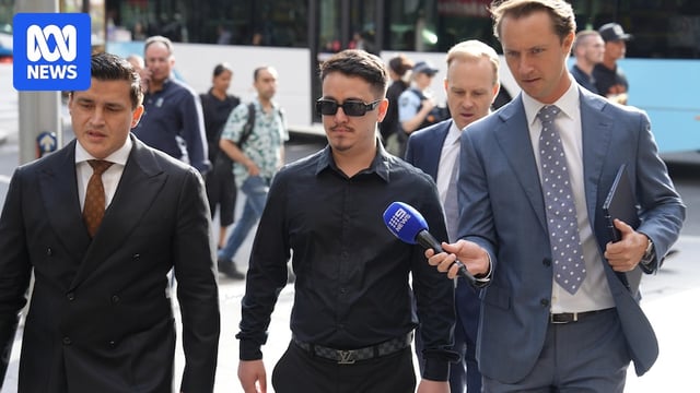 A man wearing sunglasses, a black shirt and pants walks next to two men in a suit, with one holding a microphone up to him.