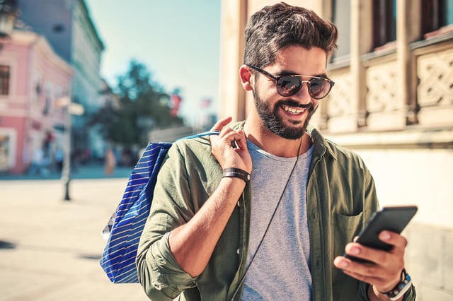 In this stock image, a man wearing sunglasses outside pauses shopping to look at something on his mobile phone.