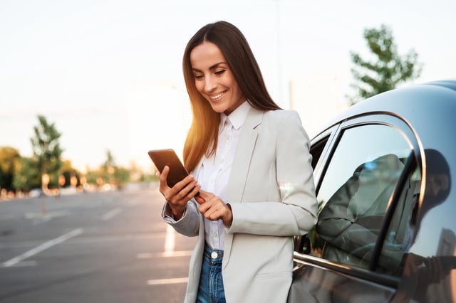 In this stock image, a woman wearing a blazer and jeans smiles while looking at her mobile phone near a modern car outdoors.