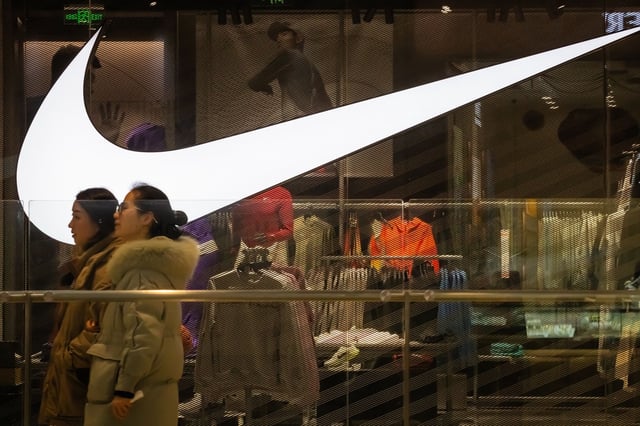 Two women walk past a Nike store featuring its large illuminated swoosh logo and interior displays of sportswear and accessories on January 18, 2025 in Chongqing, China.