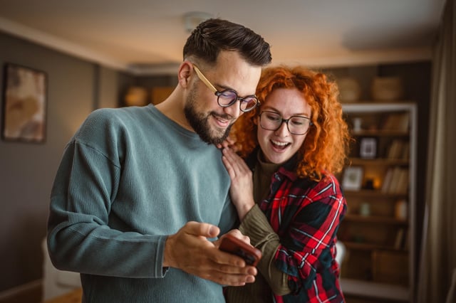 In this stock image, a couple wearing glasses looks happy while looking at a mobile phone at home.