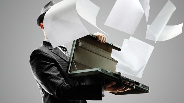 Man in dark pinstripe suit holding open a briefcase, from which papers are flying out and covering his face.