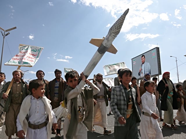 SANA'A, YEMEN - MARCH 17: Yemeni children, one of them carries a mock missile, walk next to a billboard depicting the Houthi leader Abdul Malik Al-Houthi to partake in a demonstration staged against the United States' aerial attacks on the capital Sana'a, and other provinces on March 17, 2025 in Sana'a, Yemen. Yemen's Iran-backed Houthis have declared that during the past 24 hours, they launched two missile-drone attacks on the American aircraft carrier group as retaliation for deadly US strikes targeting several of Yemen's Houth-held territories that left at least 53 people killed and more than 100 others injured, among them five children and two women. (Photo by Mohammed Hamoud/Getty Images)