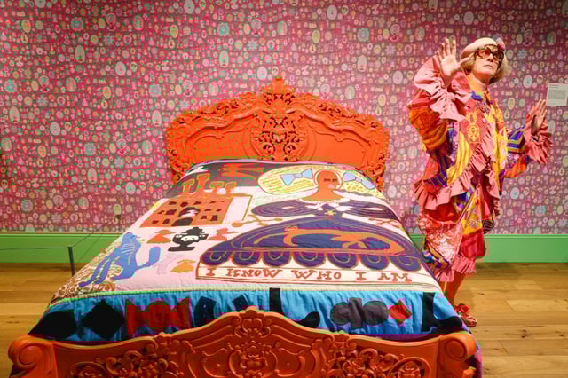 English artist Grayson Perry poses for a photograph beside "I know Who I am 2024", a cotton fabric and embroidery applique laid on a bed, during a photocall for "Grayson Perry: Delusions of Grandeur" at The Wallace Collection in London