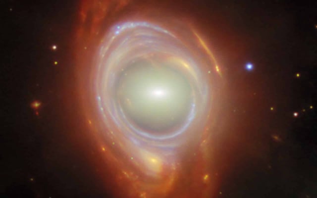 In the centre is an elliptical galaxy, seen as an oval-shaped glow around a small bright core. Around this is wrapped a broad band of light, appearing like a spiral galaxy stretched and warped into a ring, with bright blue lines drawn through it where the spiral arms have been stretched into circles. A few distant objects are visible around the ring on a black background. Credit: ESA/Webb, NASA & CSA, G. Mahler Acknowledgement: M. A. McDonald
