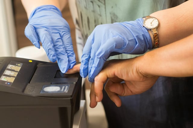 Eine Hand auf einem Fingerabdruck-Scanner – gehalten von zwei anderen Händen in Plastikhandschuhen