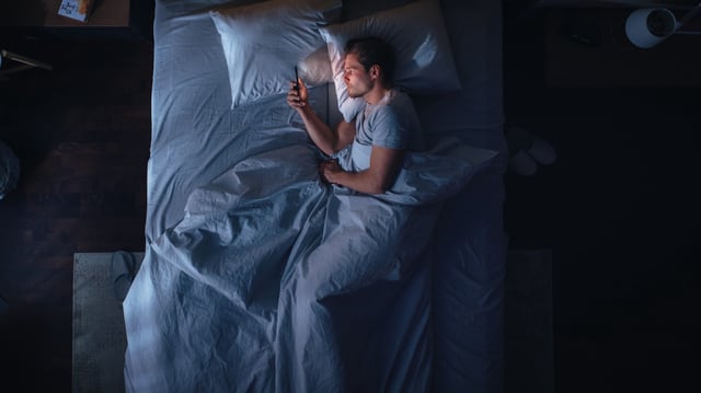 A stock image of a young man scrolling on his phone in bed at night.