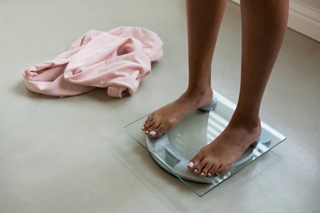 A woman stands on a scale, with her clothing on the floor beside her.
