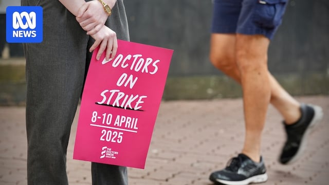 A range of doctors in red scrubs pose for photos at a strike where they hold signs.