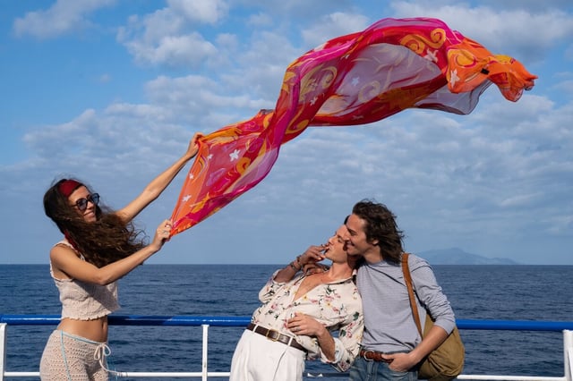 Eine Frau lässt ihr Tuch im Wind fliegen, neben ihr zwei Männer, die sich im Arm halten