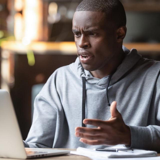 A man at a desk using a laptop and holding his hands up, while having a confused look on his face