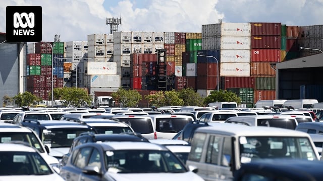 Rows of cars in a car park, with stacked shipping containers behind them.
