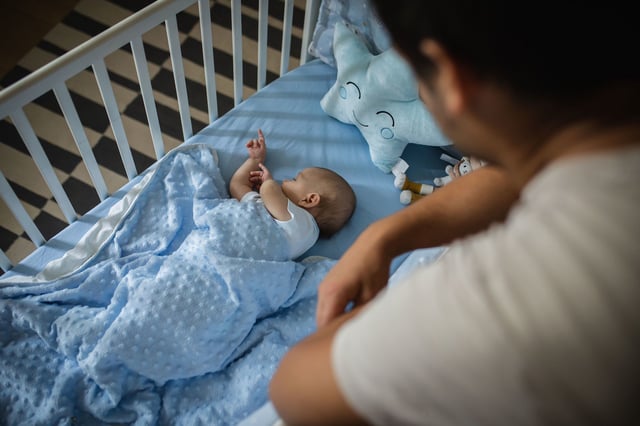 Cute silent little baby boy in white babysuite is in his crib, lying on his back, covered with blue blanket. He is sleeping while his father taking care of him. Love emotion.