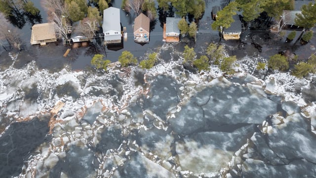 Ice forms near a row of trees near a set of houses with flooded yards, as seen from the air.