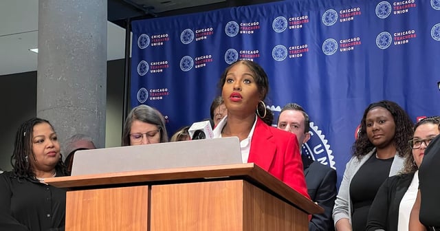 A Black woman wearing a red suit jacket speaks from behind a wooden podium and in front of a blue background an a group of people stand next to her.