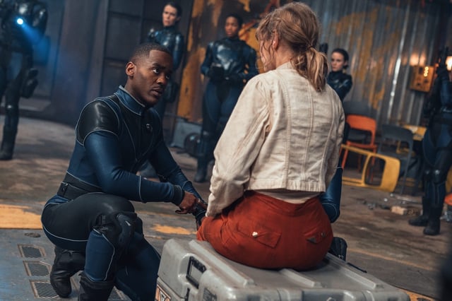 The Doctor talking to a woman sat on a box in the middle of a futuristic room.